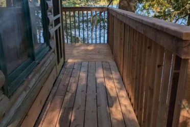 A sturdy, sunlit wooden deck and railing expertly constructed by KSC, extending from a rustic cabin towards a calm lake surrounded by lush pine trees.